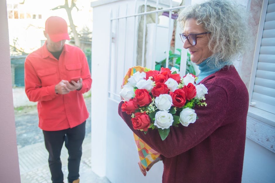A woman with curly gray hair and glasses holds a vibrant floral bouquet featuring red and white roses, wrapped in colorful paper, during a flower delivery at her doorstep. She wears a burgundy sweater
