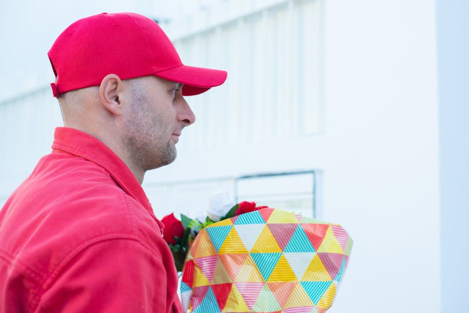 A man dressed in a red cap and red jacket is holding a large, colorful bouquet wrapped in fabric with a geometric pattern of triangles in yellow, blue, red, and green. The bouquet contains fresh flowe