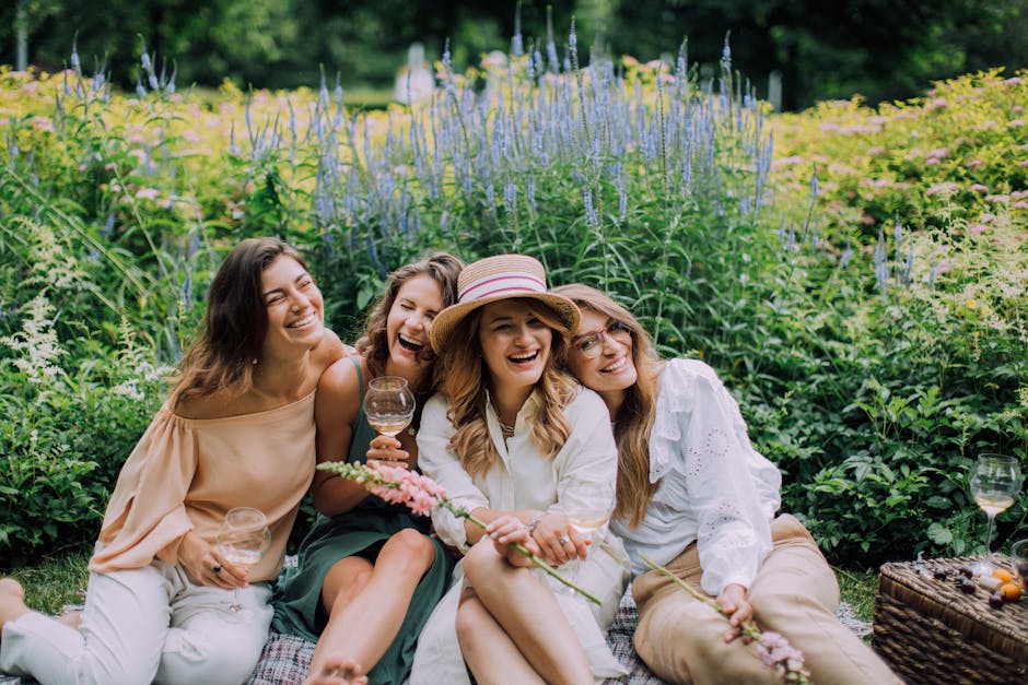 A group of four women sitting together on a picnic blanket in a lush garden setting with vibrant greenery and tall purple flowering plants in the background. They are smiling and enjoying each other's