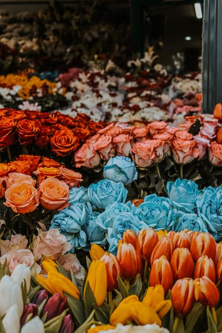 A vibrant display of fresh flowers arranged in a florist shop, featuring a variety of blooms including orange and yellow tulips in the foreground, soft pink roses, bright orange roses, and elegant blu