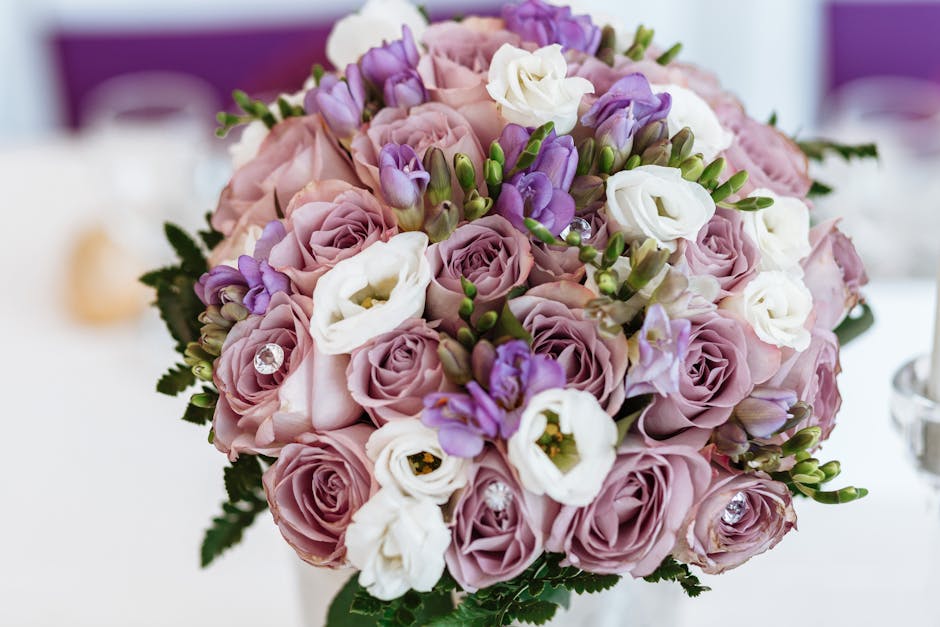 A close-up image of a floral bouquet arranged with soft pink roses, white eustoma, and purple freesia, accented by green foliage. The flowers are fresh with delicate, velvety petals and are arranged i