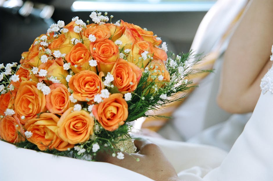 A close-up of a vibrant bouquet held by a person dressed in white, featuring closely arranged orange roses with delicate, ruffled petals, complemented by small white filler flowers and lush green foli